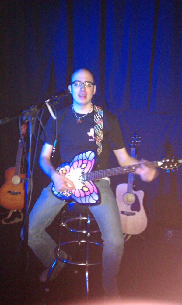 Picture of a pre-transition Eliska sitting on stage, with a butterfly guitar on, eyes closed. 