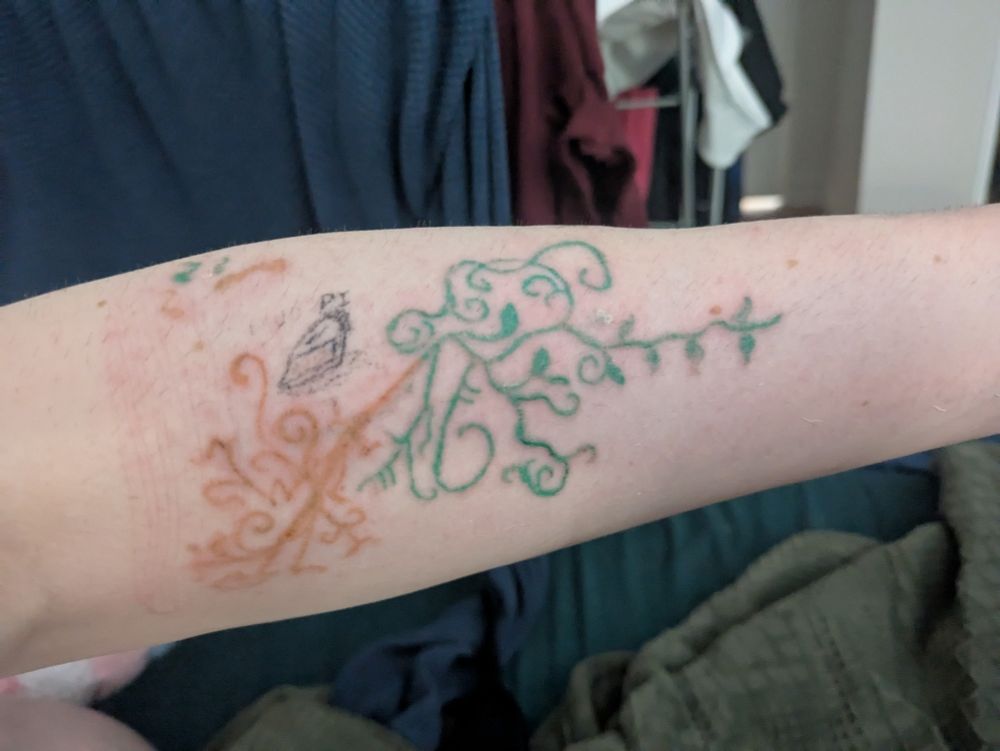 The Fall System's left arm. There is a tattoo of a sprawling vine in green ink, extending towards Fall's wrist, and a system of roots in brown ink extending toward Fall's elbow. A more faded tattoo of a slice of pie with the word "Pi" above it sets above the roots portion of the vine tattoo. 