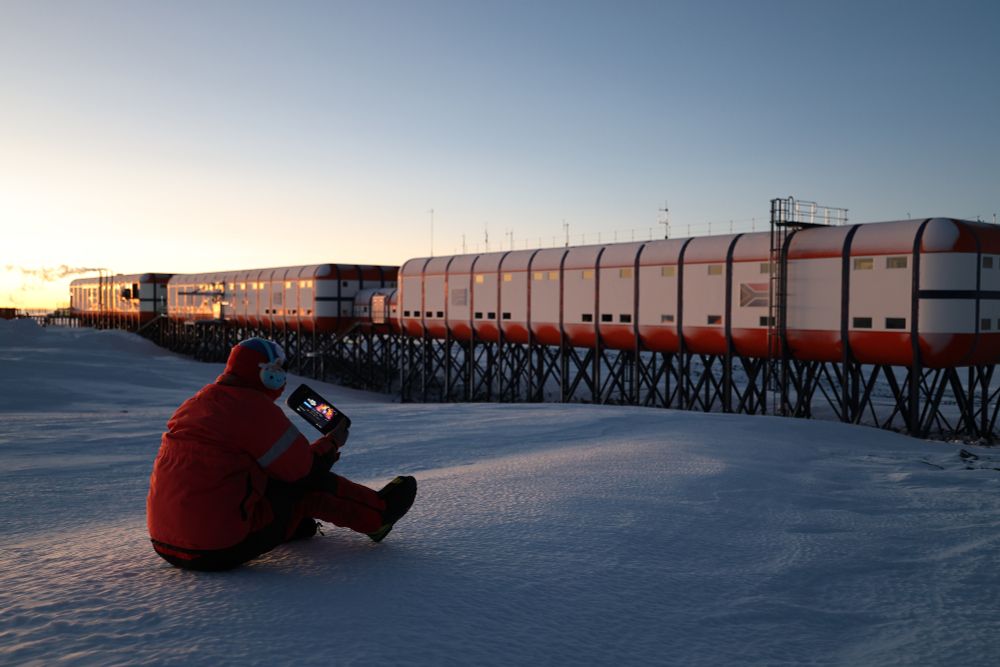 A person sitting with their back to the camera on snow in a red thermal outfit, there's a research station with a South African Flag on the side in front of them. The person is holding a steam deck with Metavoidal on the screen.
