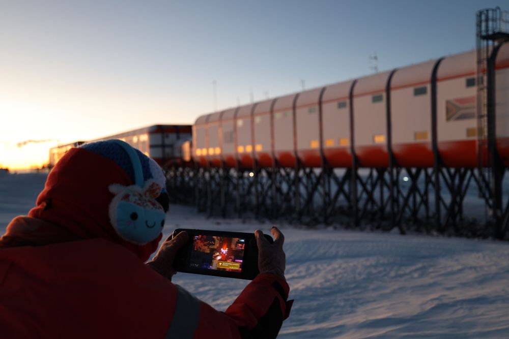 A person sitting with their back to the camera on snow in a red thermal outfit, there's a research station with a South African Flag on the side in front of them. The person is holding a steam deck with Metavoidal on the screen. They're also wearing cute earmuffs with little faces on them