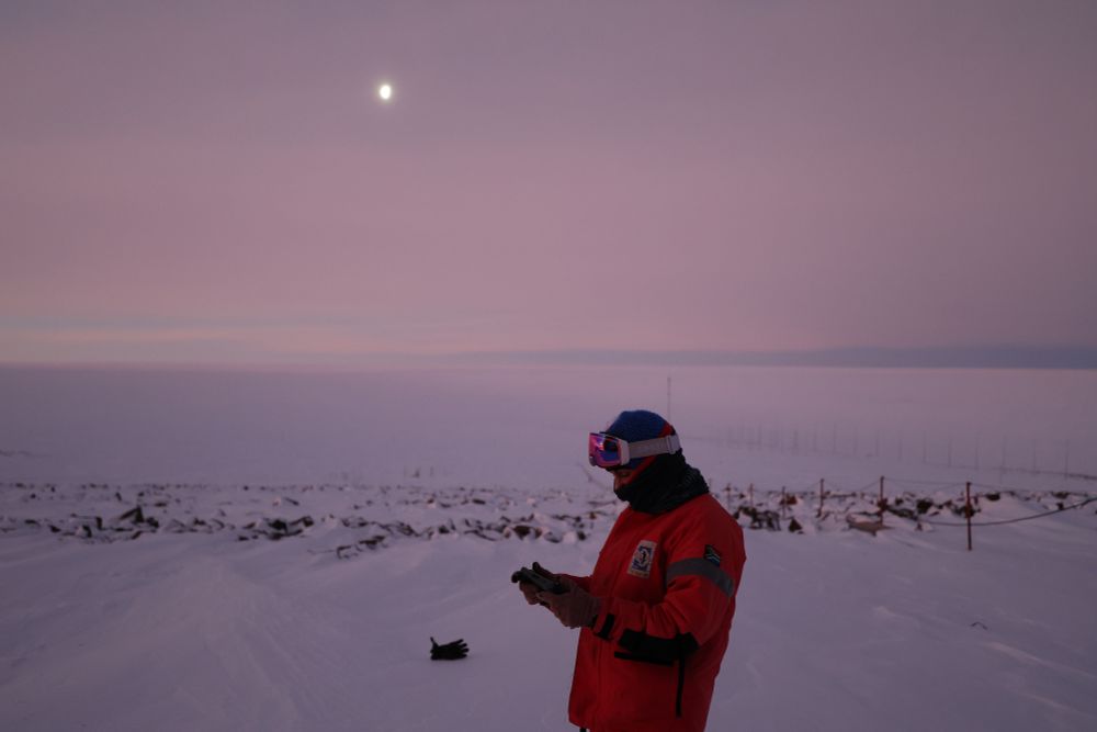 A person standing in profile in a red thermal outfit surrounded by snow,  there's nothing but white and a random glove on the floor behind them. The person is holding a steam deck with Metavoidal on the screen.