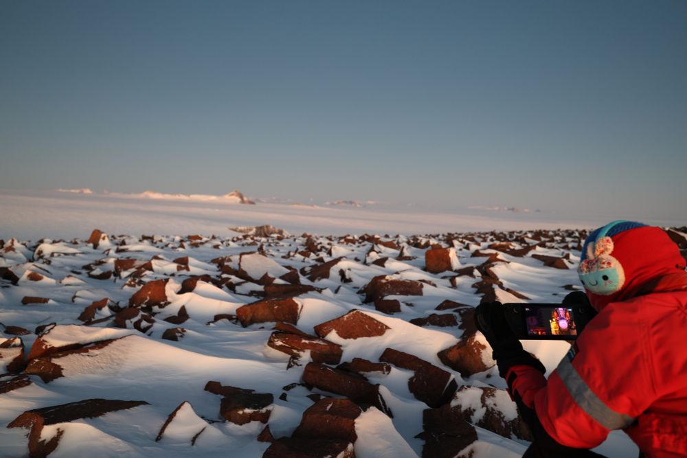 A person sitting with their back to the camera on snow in a red thermal outfit, there are a bunch of jagged rocks sticking out of the ground with snow on them. The person is holding a steam deck with Metavoidal on the screen.