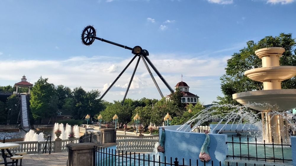 Black Widow pendulum ride at Kennywood in the distance swinging at one of its apexes, with Pittsburg Plumge spill ride in the left bottom corner and a water fountain in the bottom right.