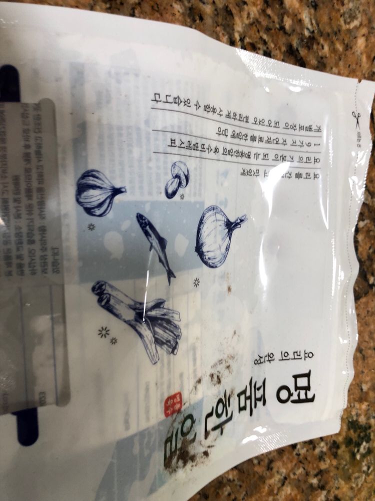Picture of a plastic bag that once contained some food, covered in Korean writing and with pictures of a mushroom, garlic, a fish, celery, and garlic.
