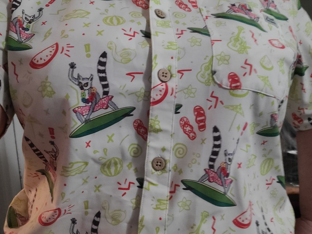 A cream-colored button-down short-sleeve camp shirt with a repeating print of a large ring-tail lemur wearing a flower lei and jam shorts, riding a surfboard. The print is accentuated with small red flip-flops, zig zags, and fruit pieces, and small lime green watermelons, fruit, spirals, and ukuleles.
