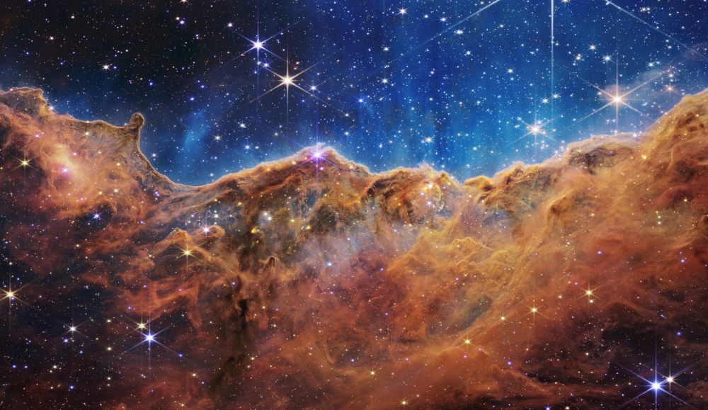 This is a NIRCam (Near-Infrared Camera) image from the James Webb Space Telescope of the "Cosmic Cliffs" in the Carina Nebula. The image is dominated by textured formations of gas and dust in warm tones of orange, brown, and gold, resembling a rugged cliff face. Bright, star-forming regions are visible within the cliffs, glowing with a soft light. The top portion of the image shows a transition to a deep blue, star-filled space. Numerous distinct stars are scattered throughout the image, some displaying a starburst effect from the telescope's optics.