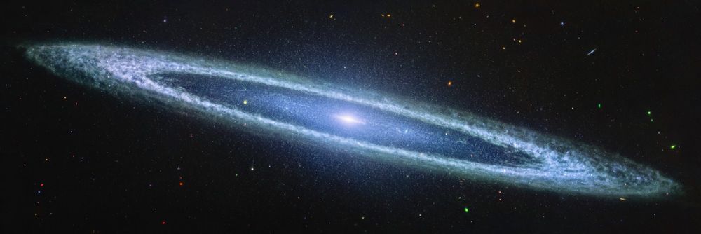 Image of a galaxy on the black background of space. The galaxy is a very oblong, blue disk that extends from left to right at an angle (from about 10 o’clock to 5 o’clock). The galaxy has a small bright core at the centre. There is an inner disk that is clearer, with speckles of stars scattered throughout. The outer disk of the galaxy is whiteish-blue, and clumpy, like clouds in the sky. There are different coloured dots, distant galaxies, speckled among the black background of space surrounding the galaxy