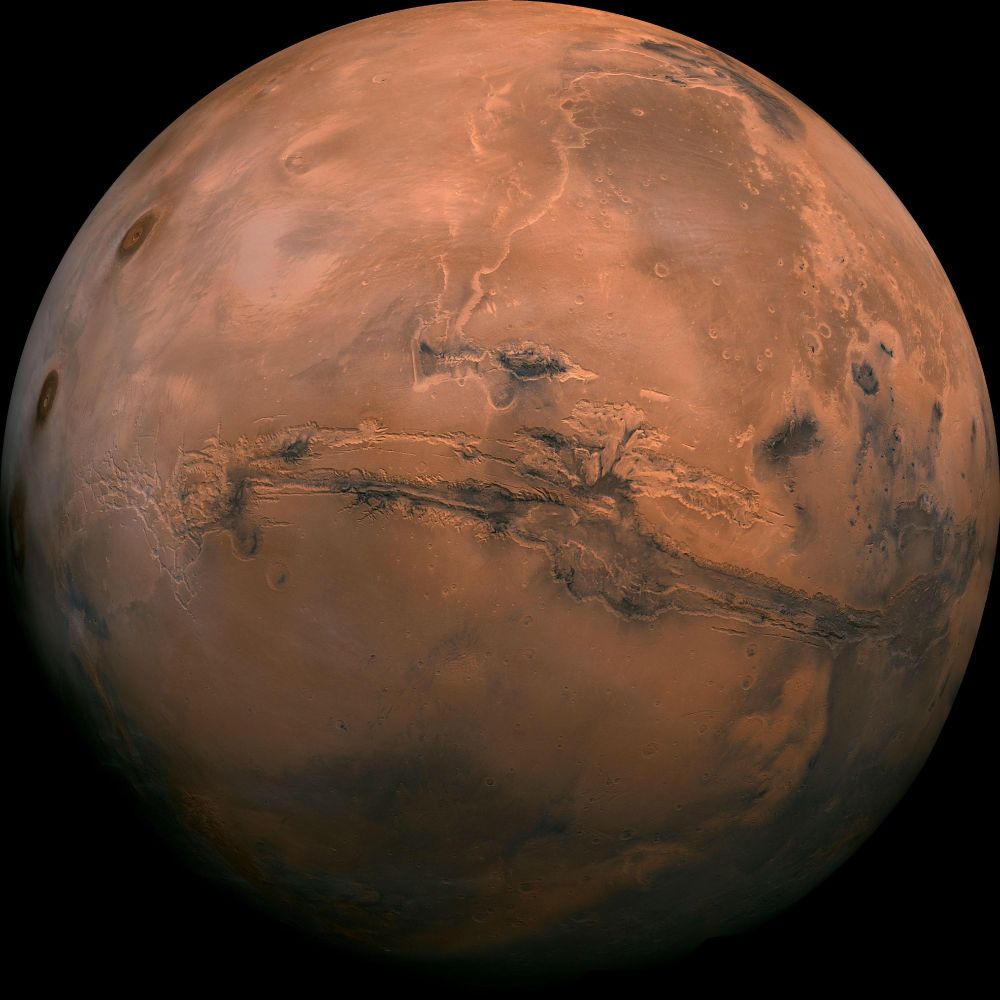 A round, rusty orange planet against the blackness of space. The surface is a mix of lighter and darker orange areas, with some darker markings that look like smudges or streaks. There's a lighter, whitish patch at the top and bottom, which are the polar ice caps.
