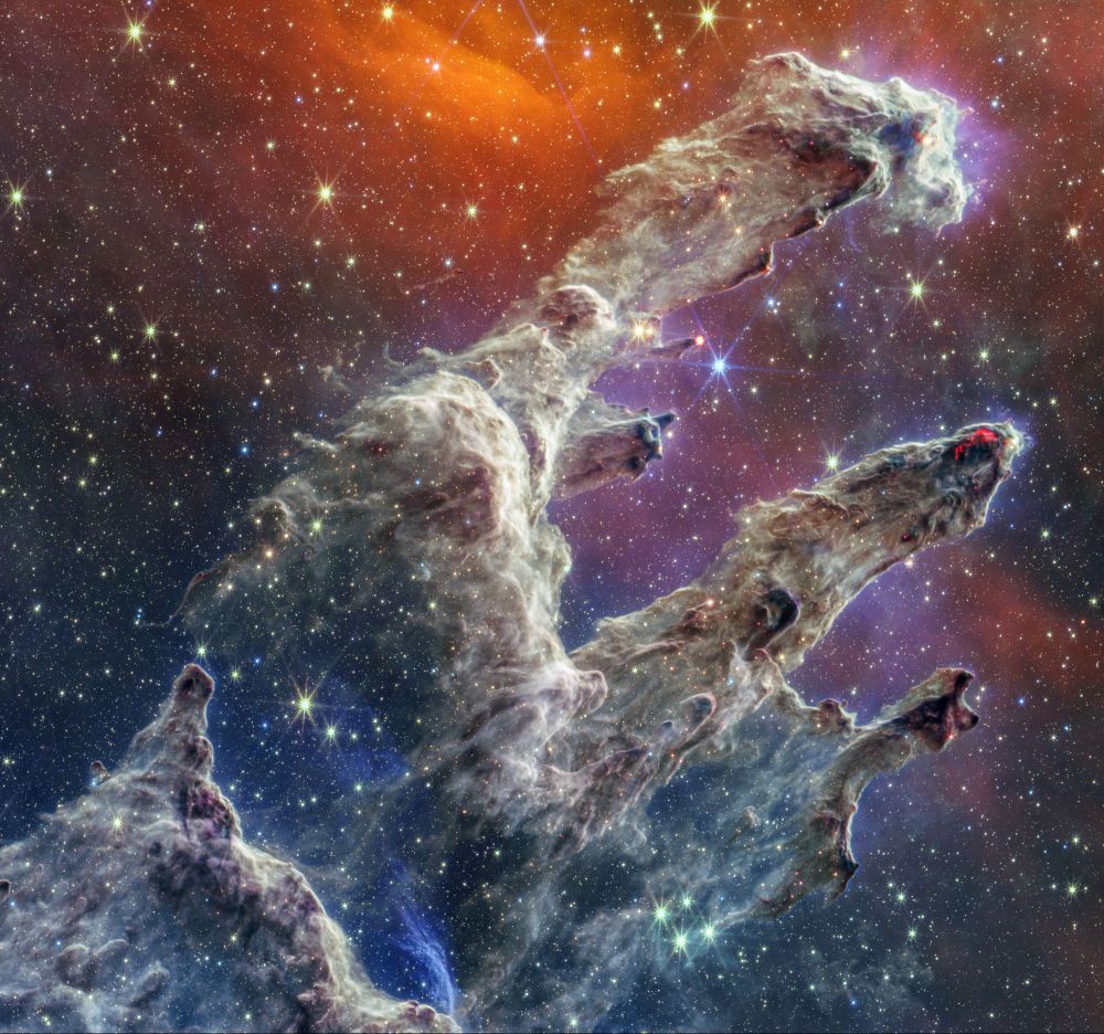 Semi-opaque layers of blue, purple, and grey gas and dust start at the bottom left and rise toward the top right. There are three prominent pillars. The left pillar is the largest and widest. The background is orange near the top and dark blue and purple near the bottom. Some blue and white stars dot the overall scene.
