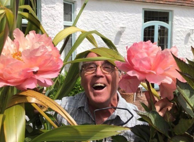 Patient contributor Kevin Coyne at home in his garden. He is peering out from between two pink flowers with a cheerful grin. 