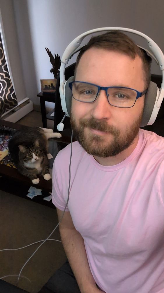 A picture of me with Kayko, my cat, laying on a glass coffee table behind me, looking in my direction. I'm wearing a headset, pink shirt and glasses. Kayko has mostly dark coloured fur with white paws and a white streak from his forehead down his chest 