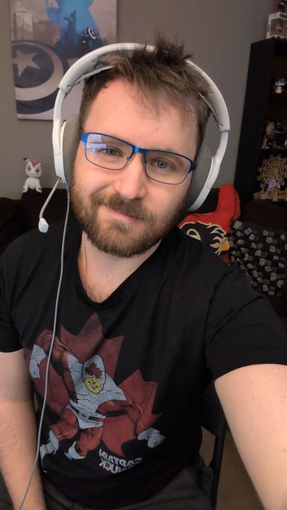 A picture of me in my living room wearing a black t-shirt with am image of Captain Canuck on it. I'm wearing a white headset and glasses with a blue frame. In the background is a canvas of captain America, a Gomamon plushie, a pillow shaped like a foam finger for the Calgary Flames and a shelf with various objects on it