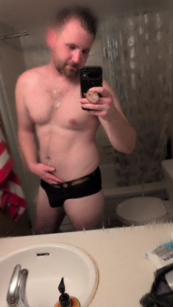A picture of me in my bathroom wearing black briefs with gold writing. 