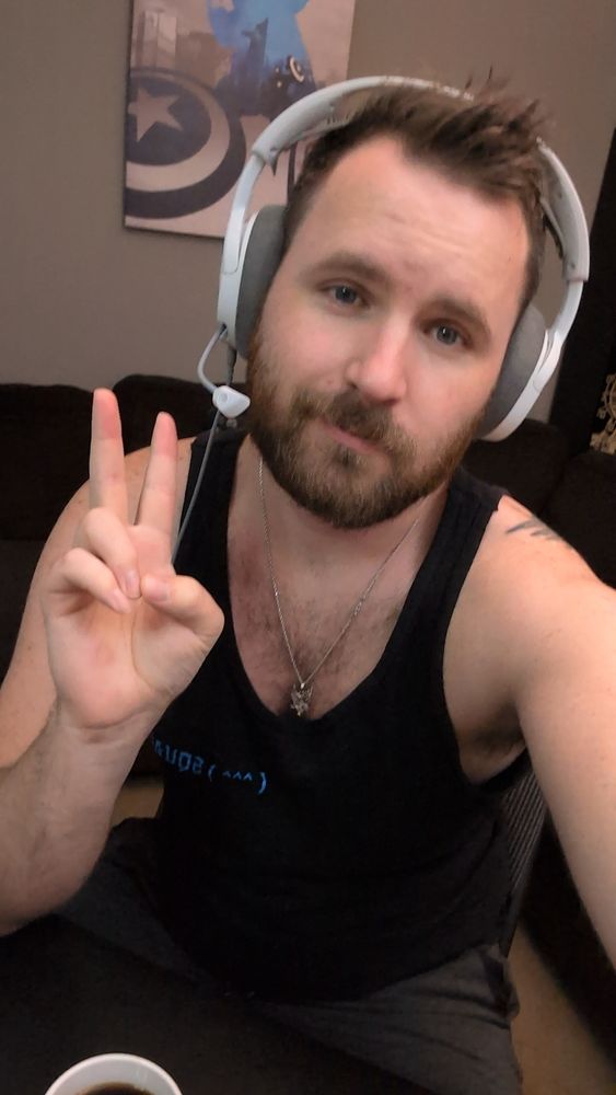 A picture of me wearing a black tank top and white headphones. I have a beard and a silver chain. My hand is giving a peace sign and there is a cup with coffee infront of me and a captain America canvas on the wall behind me.