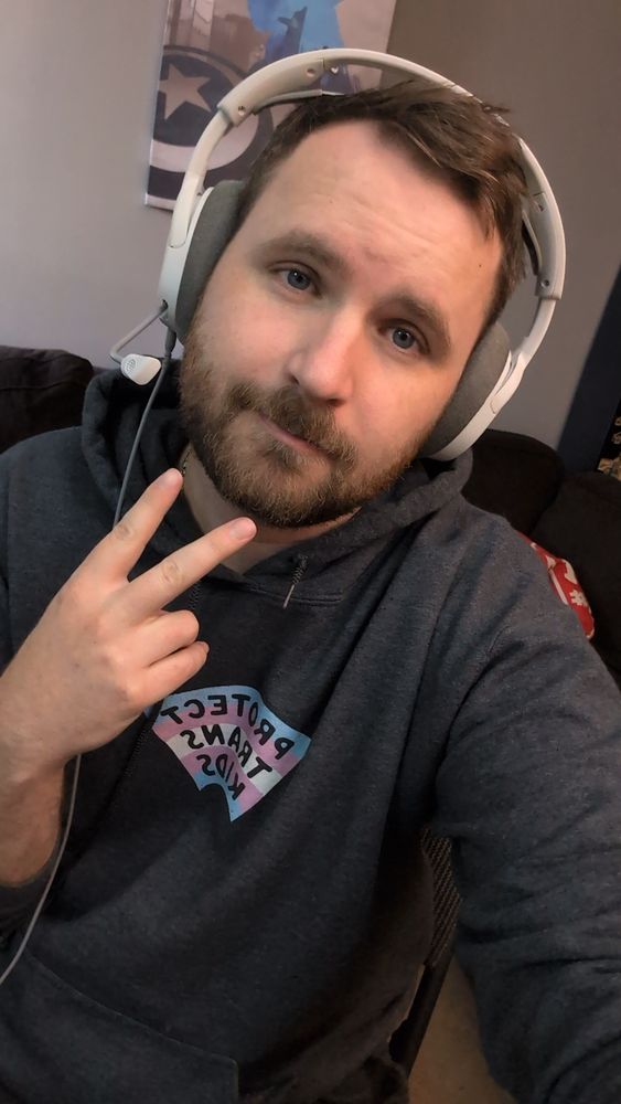 A picture of me in my living room wearing a sweater that says "protect trans kids" on it with the trans flag. I'm wearing a headset and there's a captain America canvas on the wall behind me 