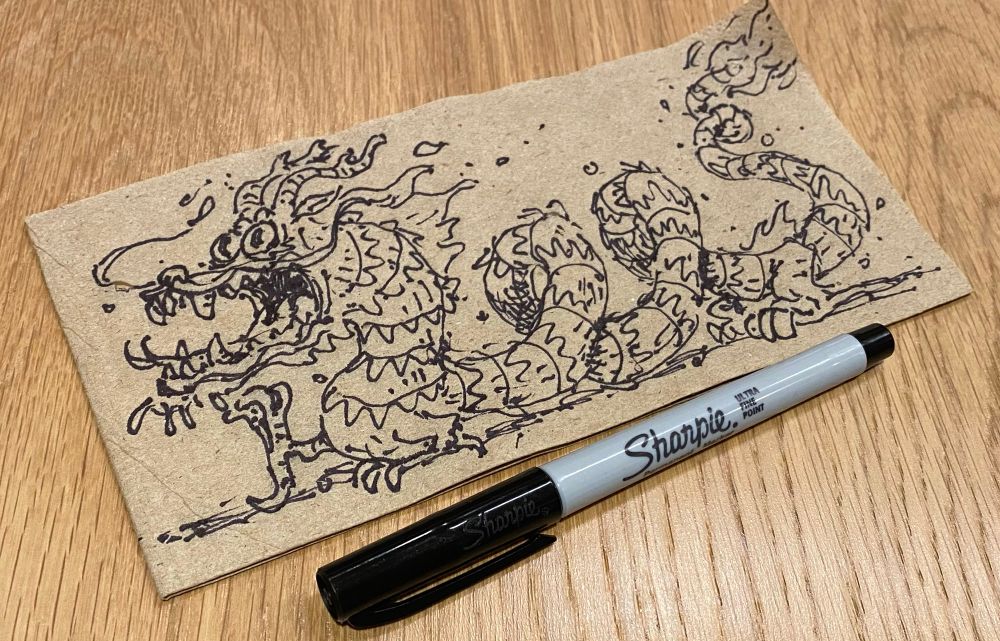 Sketch of a Chinese dragon roughly drawn on a brown napkin slightly askew with a fine point sharpie just below the napkin also at a rakish angle. 