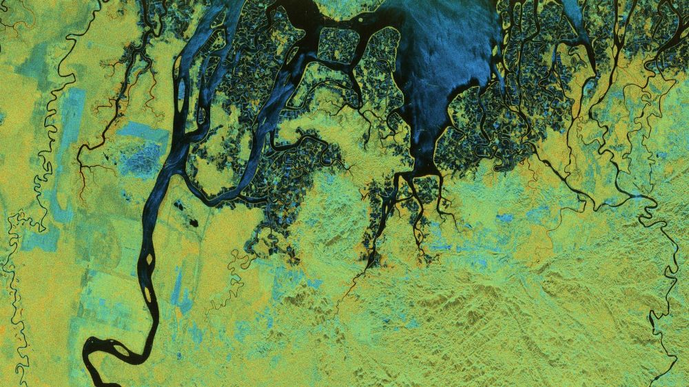 A map with the land covered vibrant shades of green, in mixed with red  in some areas and showing topographical features. The upper half shows a coastline with water, in shades of blue so dark they seem black in places. Multiple rivers snake across the landscape.