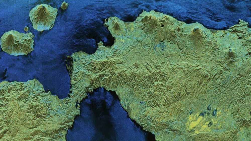 This image shows the sea, in deep shades of green, around landmasses in a wide variety of shades of vibrant green. Conical mountains pierce upwards, creating a sense of great elevation differences beneath the forest.