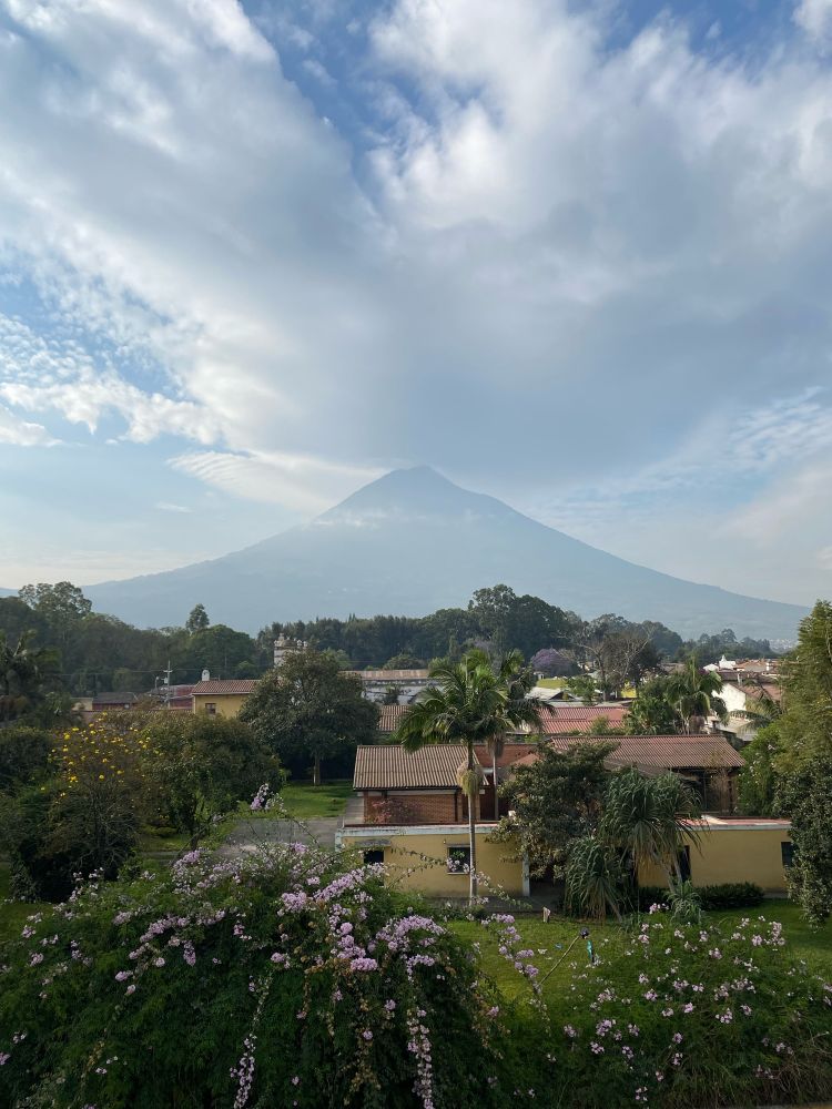 Agua, a large stratovolcano in the back of the scene, standing tall between some thin clouds. To the front, a somewhat luxurious neighborhood in Antigua Guatemala, with a lot of beautiful gardens and colonial style houses.