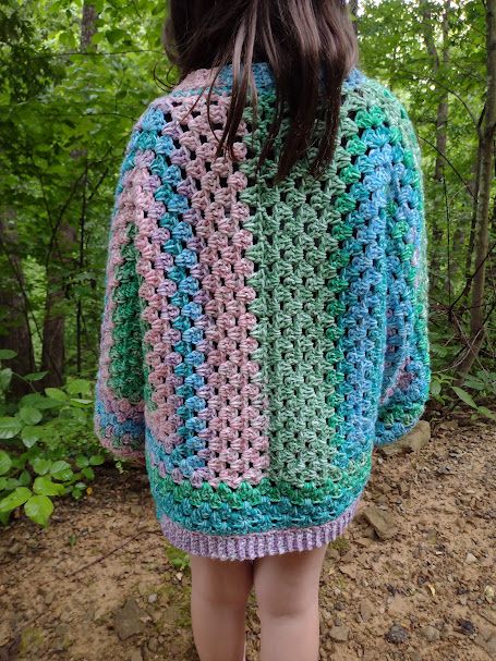 A child standing outdoors, shown from behind, wearing a hexagon cardigan crocheted by hand in pink, blue and green.