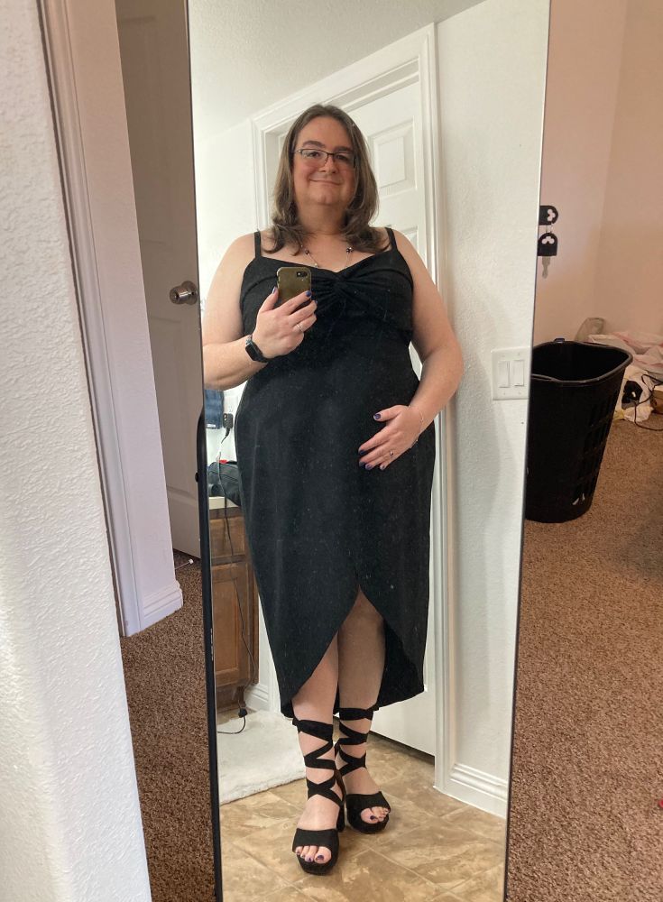 A selfie of Josie reflected in a full-length mirror. She is wearing a black dress that hugs her waist and hips and strappy sandals. 