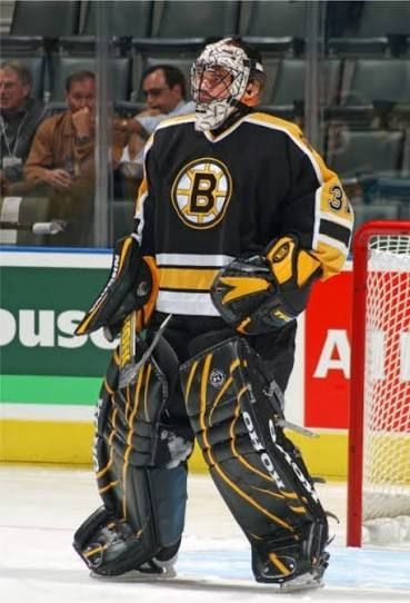A goalie standing tall in net wearing a black late 90s Boston Bruins jersey and a Gerry Cheevers tribute mask.