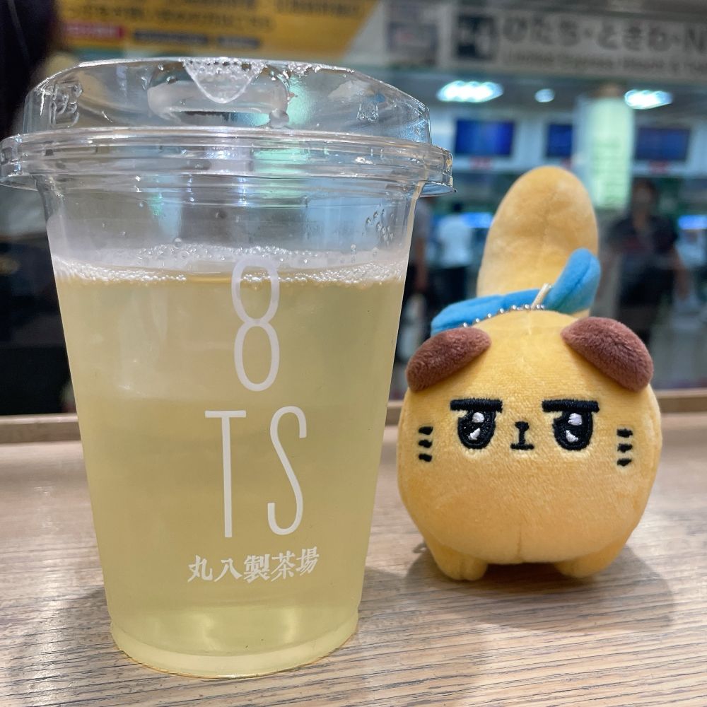 画像左から机の上に透明なカップに入ったお茶(玄米茶)、東京ばにゃ奈のぬいぐるみストラップ。
背景は駅の構内。