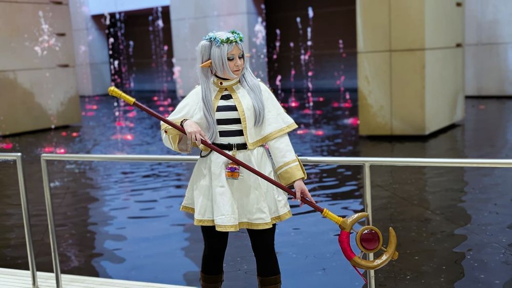 A Frieren cosplayer standing in front of an indoor fountain, spraying water from red light, she is looking downward