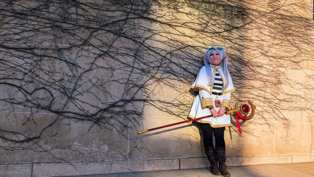 Frieren cosplayer against a sunlight stone wall covered in black ivy, a positive expression on her face 