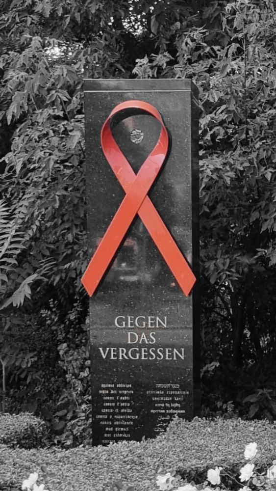 Schwarz-weiß Bild, ein großer Marmor Stein steht vor Bäumen, davor Blumen. Auf dem Stein ist eine rote Schleife, darunter steht "Gegen das Vergessen". 