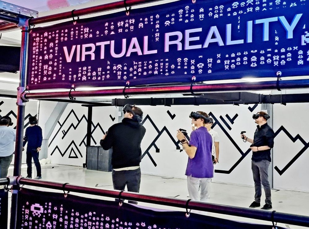 In einer Halle stehen mehrere Menschen mit VR Brillen und schauen nach oben oder seitlich. Der Bereich ist mit einer Abgrenzung versehen, auf der steht in Großbuchstaben VIRTUAL REALITY.