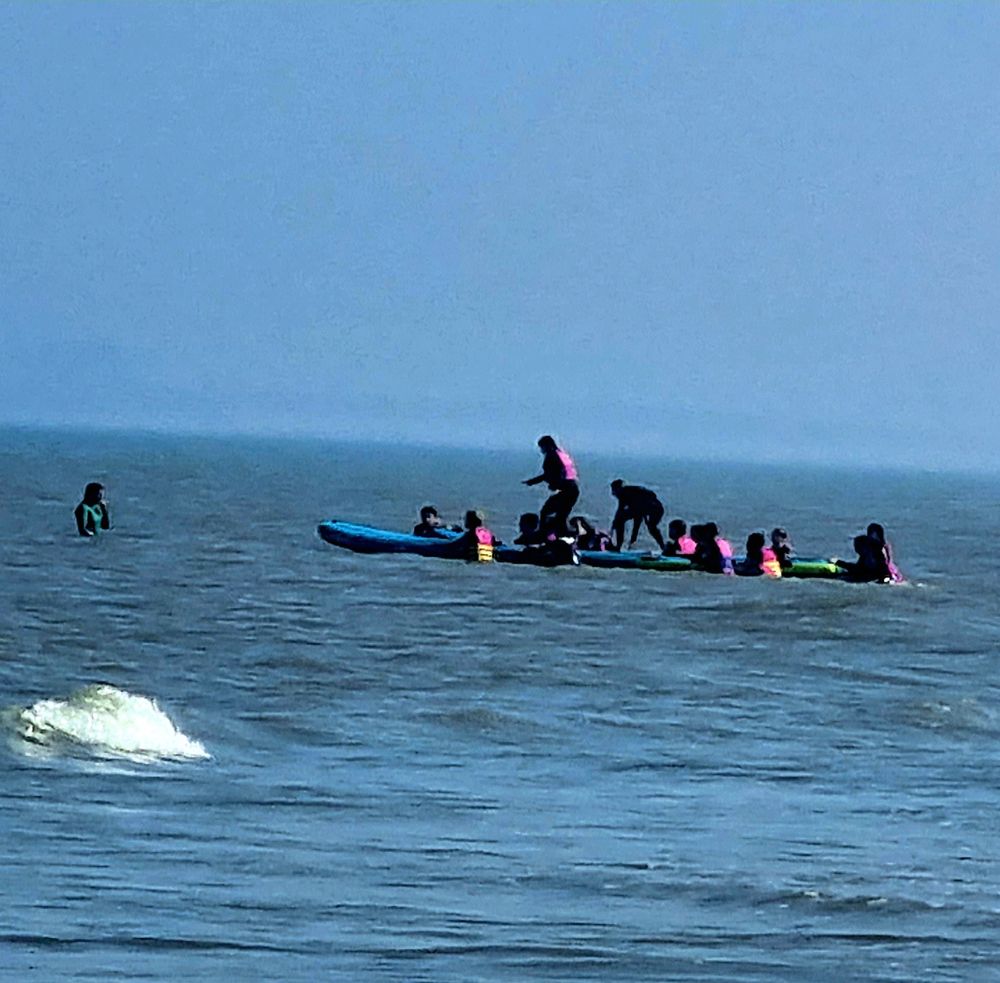 Im Meer sieht man ca ein dutzend Menschen mit Rettungswesten, das Wasser geht allen bis zum Bauch. In der Mitte der halten die Menschen zwei Boards hintereinander, auf denen zwei Menschen drüberlaufen und versuchen, Gleichgewicht zu halten und nicht ins Wasser zu fallen. 