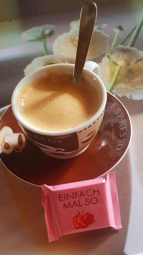 Eine Tasse mit Kaffee steht auf einem Unterteller, an dem eine kleine Schokolade angelehnt ist mit der Aufschrift "einfach mal so".