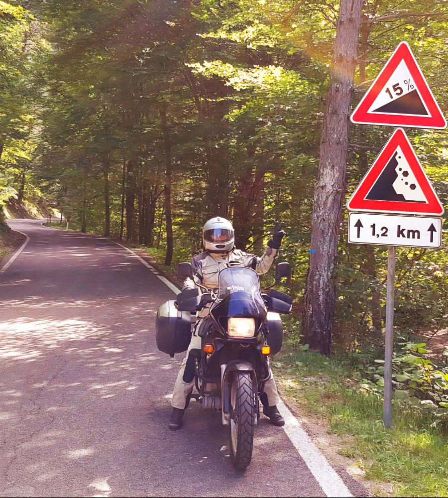 Eine kleine Straße durch einen Wald, am Straßenrand Schilder dass Gefahr durch Steinschlag und 15% Steigung. Auf der Straße ein schwarzrs Motorrad mit seitlichen Koffern, eine Person in weisser Motorrad Kleidung und hellem Helm streckt einen Arm aus zum Gruß. 