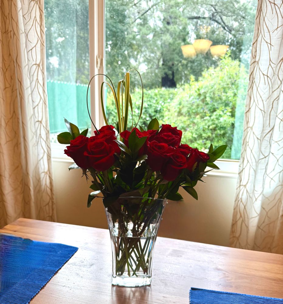 A dozen red roses in a fluted glass vase on a wooden kitchen table in front of a window. 