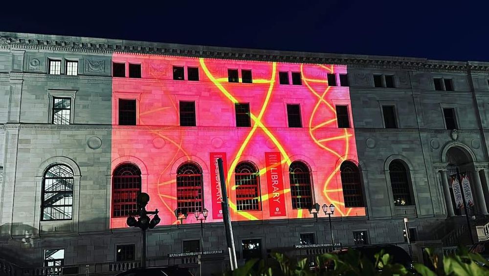 Image of an old three story brick building with many large windows a banner stating the word library. Over the face of the building, an animation of a DNA double helix illuminates the space.