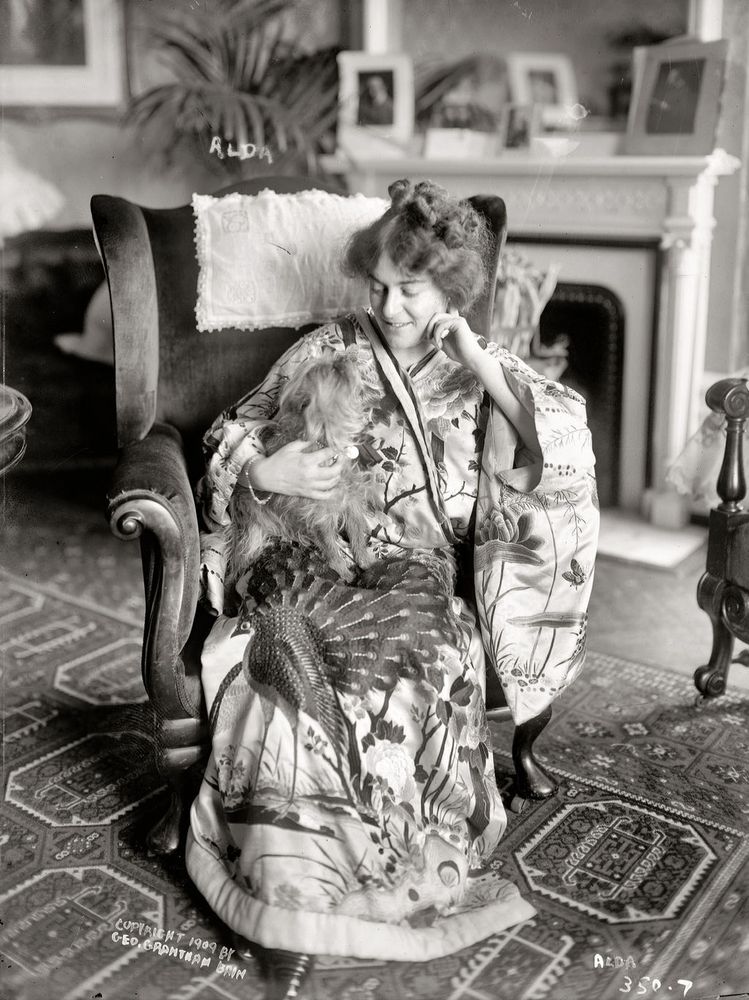 B&W photo of a lovely woman wearing an elaborate robe with her hair in an updo. She's smiling down at a Yorkshire-terrier-like lapdog who is gazing up at her excitedly