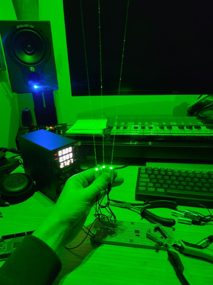 A hand holding three small green lasers above a desk. The lasers are lighting the whole room up in green. The desk is covered in several tools, a breadboard and a bench power supply.