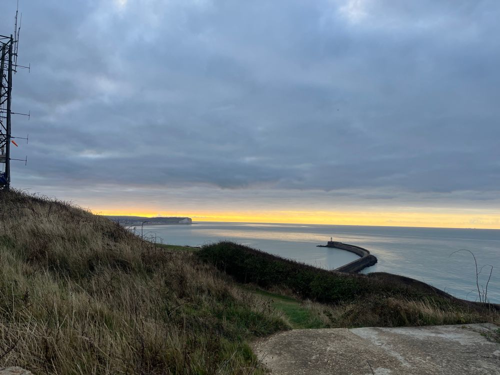 Closer shot of the sunrise behind the lighthouse, with the sun peaking over the cliffs in the distance and making parts of the sea’s surface shimmer 