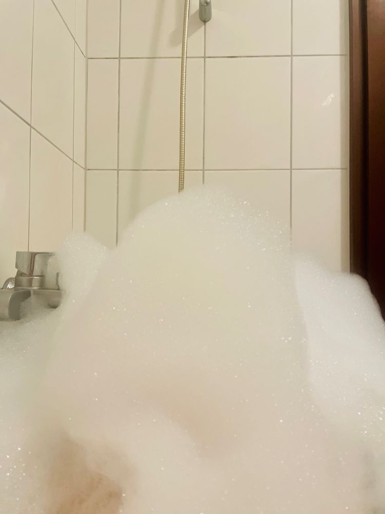 Ein Turm aus Badeschaum in einer Badewanne vor einer weißen Fliesenwand, der in der Höhe über den Wasserhahn reicht