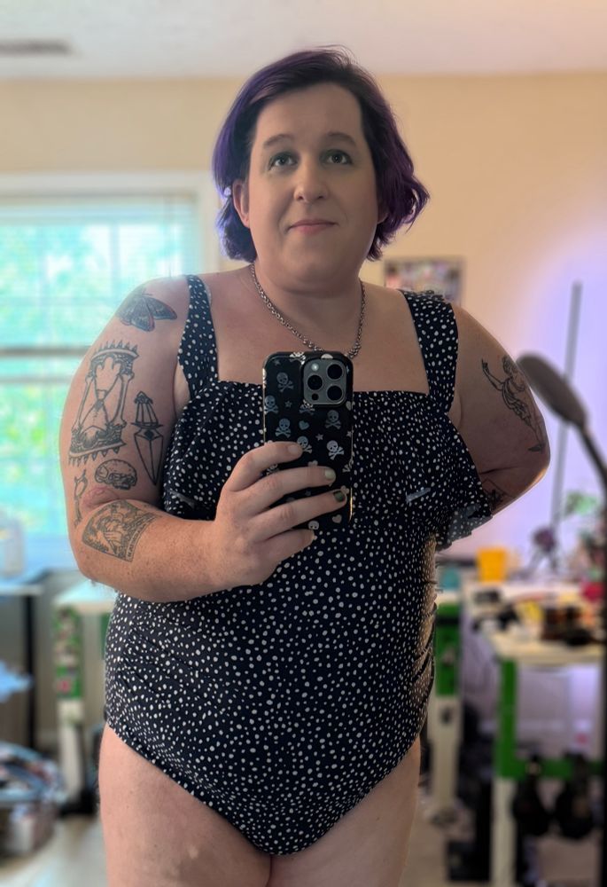 A woman with purple hair and tattoos wearing a one piece swimsuit. The swimsuit is black with irregular white dots 