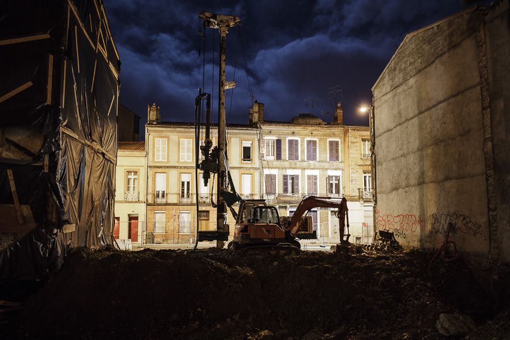 Scène de nuit : une pelleteuse sur les ruines d'un bâtiment récemment démoli entre deux bâtiments encore en place. De l'autre côté de la rue, les bâtiments sont baignés dans la lumière d'un lampadaire. Le premier plan et les bâtiments sur les côtés sont dans l'ombre. La pelleteuse émerge au milieu de l'image.

Quartier Belfort à Toulouse. Angelos Detsis, 2014