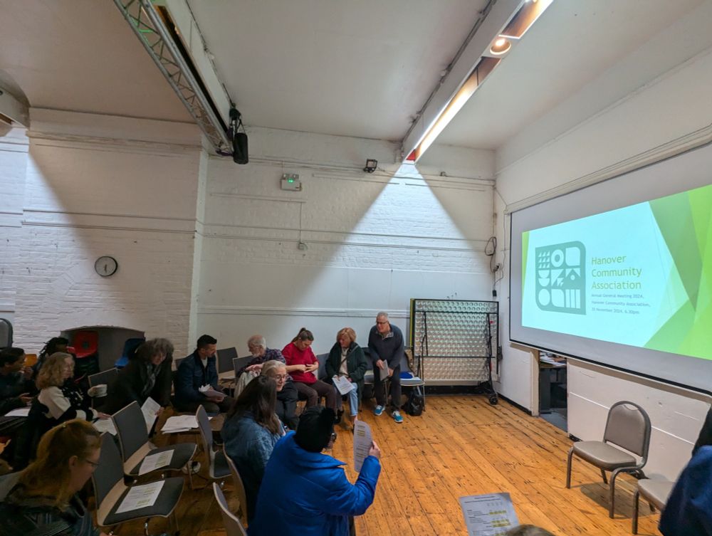 seated crowd file into Hanover Community Centre, Brighton for Annual General Meeting. Screen to right side of picture showing Hanover Community Association General Annual General Meeting 2024, 25 November 2024, 6.30pm