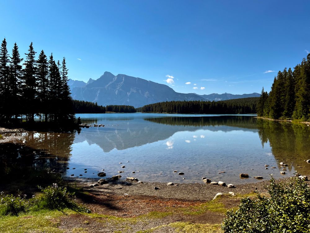 A still mountain lake reflects the mountains in the background, a small island on the left hand side with pine trees, the pine trees that line the shore on the right side and the blue sky that is peppered with a few white fluffy clouds. 