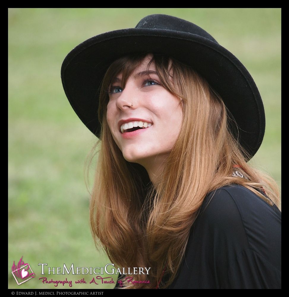 A high-quality photograph captures a young woman with fair skin, light brown hair, and blue eyes, wearing a black wide-brimmed hat and a black top. She smiles warmly, with a nose ring on her left nostril. The background is a blurred, green outdoor setting. The lighting is natural, enhancing her features and creating a pleasant, relaxed atmosphere.