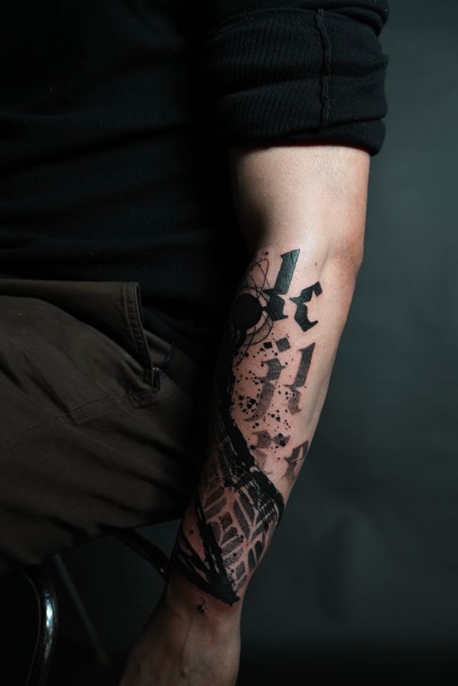 Abstract tattoo on the outer, lower forearm featuring distorted, glitchy old English lettering, art deco floral patterns, grungy brush strokes and a solid black circle with erratic circular and elliptical sketch lines orbiting it