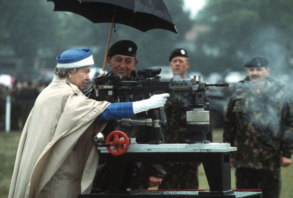 Queen Elizabeth firing a round from an assault rifle locked into shooting position on a table. A bemused SAS member looks on as he holds her umbrella
