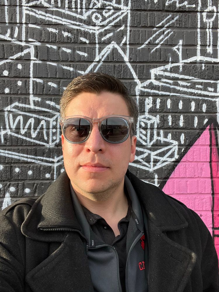 Selfie of me wearing sunglasses and standing in front of a mural of a cityscape painted in white on a black wall