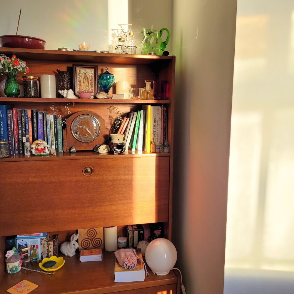 A retro drinks cabinet which instead holds occult books, tarot decks, and an altar to Santa Muerte. The morning sun is glowing.