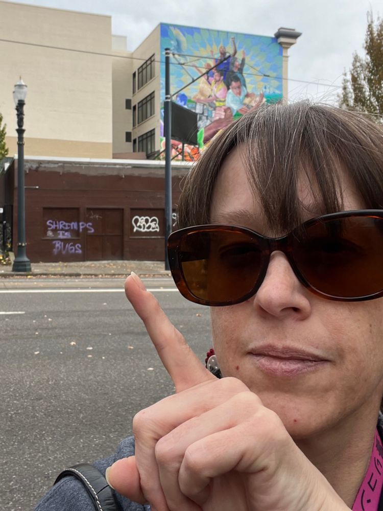 Selfie of a frumpy, middle-aged, white woman pointing at graffiti on a wall behind her. The wall is brown, the tag is lavender.